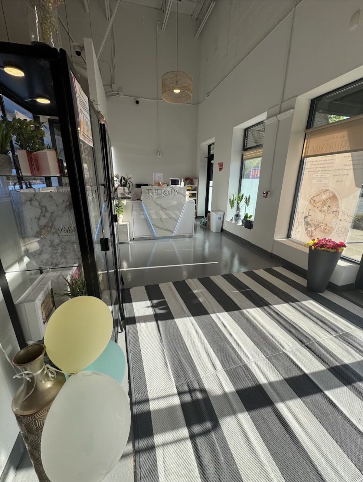 The salon entrance. A black and white striped rug leads past glass display cabinets to the white marble front desk. Sunlight comes in through the front windows.
