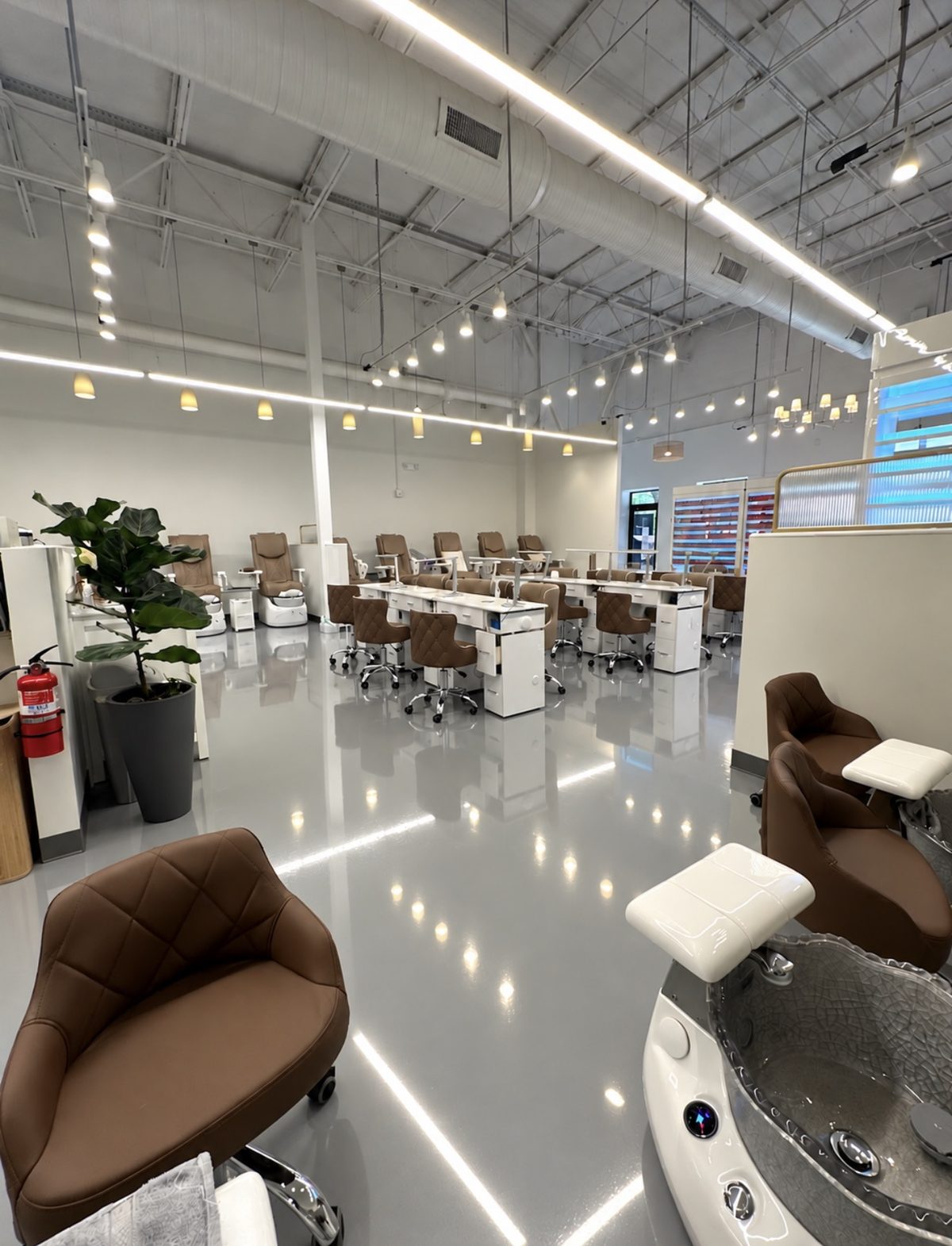 Rows of white manicure tables with brown chairs, and pedicure chairs along the far wall, under a high white ceiling with many small lights.