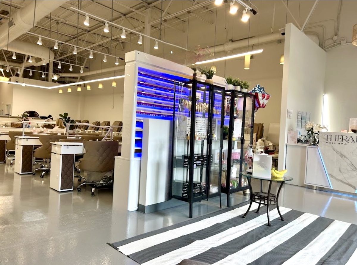 A lit wall of nail polish colours beside black glass display cabinets, with the manicure stations behind and the marble front desk at the right.