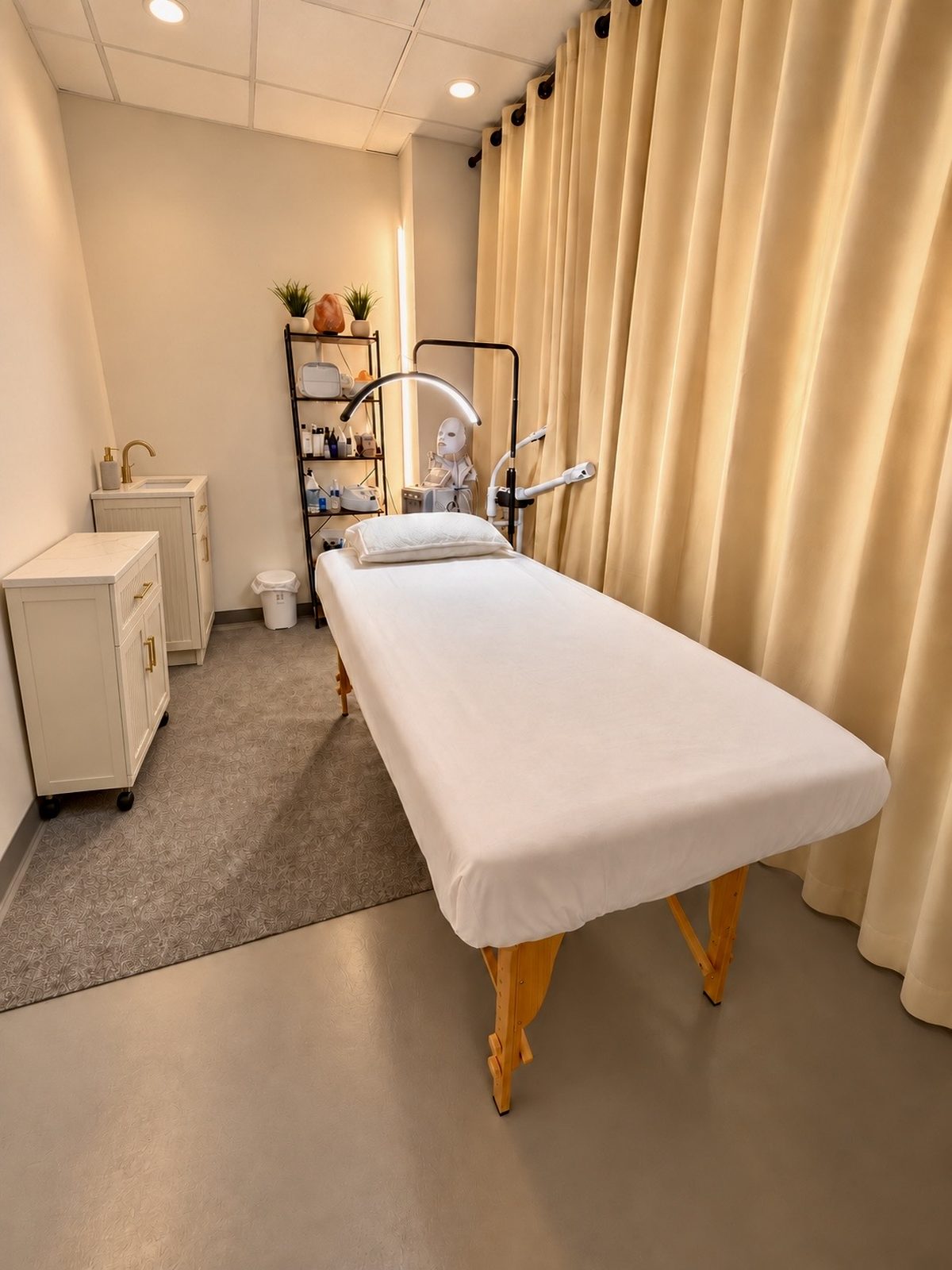 The skin care room: a treatment bed with a white cover and pillow, a facial steamer and lamp, a small sink, and warm cream curtains.